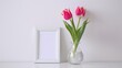 A close up shot captures a lovely tulip flower displayed in a glass vase surrounded by a picture frame set on a white wooden table against a clean wall at home This charming scene embodies 