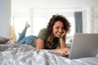 Cheerful smiling curly haired young woman using laptop for video call while lying on cozy bed at home.