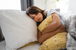 Sorrowful depressed young woman with closed eyes hugging pillow in a bed.