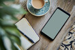Mockup of smartphone with blank screen, coffee cup, open paper notebook and green plant leaves on wooden table. Top view