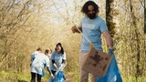 Fototapeta Tulipany - Man activist using tongs to grab garbage and plastic waste, picking up trash and cleaning the forest area. Volunteer sorting rubbish and recycling it, preserving the nature. Camera A.