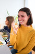 Vertical. Pretty young Caucasian woman smiling holding glass white wine sitting at table at unfocused family meal in background. Generations together enjoying themselves at a celebratory event