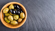 Black and green olives and oil in wooden bowl on black slate background. Top view with space for text.