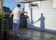 Man cleaning wooden deck with hand-held pressure sprayer, shadow on the wall in the afternoon sun.