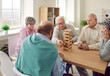 Group of elderly people playing board games in senior care centre or retirement home. Several old men and women sitting around table and taking turns to take blocks out of wooden tower