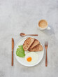 Breakfast with fried egg, avocado and toasts on white plate, bronze colored coffee cup and silverware on gray table with copy space. Top view.