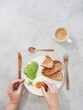 Top view of woman hands eating breakfast with fried egg, avocado, toasts and coffee on grey background.