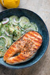 Blue bowl with grilled salmon steak and cucumber salad, vertical shot on a beige granite surface, middle close-up, selective focus