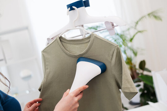 Cropped woman using steaming iron to ironing fashion shirt at home in bright cozy room. Female holding electric steamer, using modern technologies