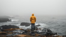   Person In Yellow Jacket Stands On Rocky Cliff, Gazing At Foggy Ocean