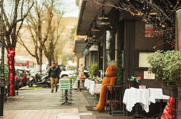 A cozy teddy bear sits on a wooden table outside a charming restaurant, surrounded by the bustle of a lively street