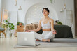 Yoga mindfulness meditation online. Woman practicing yoga with online lessons in laptop at home. Woman sitting in lotus pose meditating relaxing indoor. Girl doing breathing practice. Yoga at home