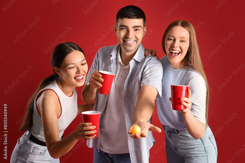 Young friends with cups and ball for beer pong on red background
