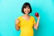 © luismolinero - Pregnant woman holding an apple isolated on blue background pointing to the side to present a product