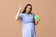 Pregnant young woman with small electric fan showing OK gesture on beige background