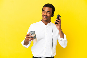 Wall Mural - African American Telemarketer man working with a headset over isolated yellow background holding coffee to take away and a mobile