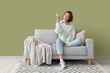 Smiling young woman with air conditioner remote control sitting on sofa near green wall