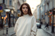 Mockup. Young woman wearing blank white crewneck sweatshirt. Young girl posing against blurred European city street in sunny day. Mock up template for sweatshirt design, print area for logo or design