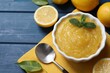 Delicious lemon curd in bowl, fresh citrus fruits and spoon on blue wooden table, above view