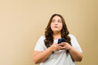 © AntonioDiaz - Thoughtful plus-size woman holding a phone, gazing towarads copy space in a studio setting
