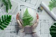Top view of an engineer holding a white helmet with a green fern leaf on a blueprint construction plan against a background of a sustainable building concept.
