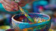 A beginners hand dips a delicate paintbrush into a jar of glaze carefully applying colorful designs onto a clay bowl.