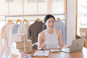 Wall Mural - Beautiful Asian female fashion designers or dressmakers show the sample clothes to the client in the online meeting. Fashion designer working in the studio. Small business concept