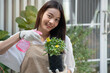 Beautiful young woman spends her weekends tending to the plants in her garden, spraying beautiful flowering plants with water sprayer at home, looking at camera