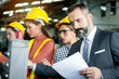 © Stella - Senior businessman reading work information from document paper at manufacturing plant factory with industrial engineer workers with safety helmet as blurred background. Manager inspects workplace.