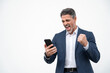 © Stock 4 You - Happy delighted satisfied business man looking at mobile phone screen gesturing yes winning pose, isolated background. Overjoyed excited businessman in suit make winner gesture reading happy good news