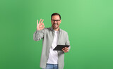 Fototapeta Panele - Portrait of young man showing OK sign and checking social media apps over tablet on green background