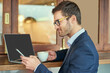 © peopleimages.com - Businessman, laptop and phone in cafe with glasses for remote work with reading sms, work break and social media chat. Male lawyer, drink and computer in coffee shop for online advice and mobile text