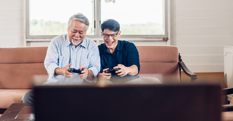 Wall Mural - Portrait of happy love asian family senior mature father and young adult son having fun enjoying holding gamepad controller play console video game together at home.