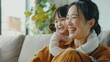 © Oulaphone - Asian woman sitting on the sofa with her daughter into hugging mom's shoulder, both smiling happy on living room.