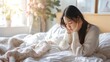 © Oulaphone - Tired asian woman sitting on bed with head in hands, feeling sick and unwell at home. Young girl suffering from falling ill or having handsome pain while alone during morning time in bedroom.