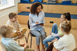© Seventyfour - High angle view at children sitting in circle during support group meeting in school with young female therapist listening