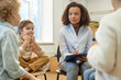 © Seventyfour - Portrait of Black young woman as child psychiatrist listening to kids during support group meeting and holding clipboard