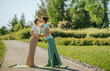 Two women in yoga attire share a joyful kiss on a sunny pathway surrounded by lush greenery. Warm, intimate moment captured in natural outdoor setting