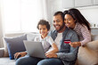 © Prostock-studio - African American family seated on couch, is making an online purchase using laptop and credit card. The parents are guiding son through the process of inputting payment information on the screen.