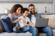 © Prostock-studio - African American family is seated on a couch, all attentively looking at a laptop screen in a living room setting. The laptop is open, displaying a bright screen with various images and text visible