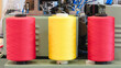 Three different colored spools of thread are on a machine. The spools are red, yellow, and orange