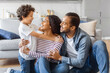 © Prostock-studio - African American man, woman, and child are seated on the floor in front of a couch. They appear to be engaged in conversation or possibly watching something together.