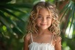 Happy smiling little blonde curly hair girl in white summer dress walking in summer tropical beach among palm leaves