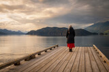 Fototapeta Most - Senior woman gazing out over a lake at sunset from a boat dock. Seen at Harrison Lake, British Columbia, with dramatic clouds and calm waters.