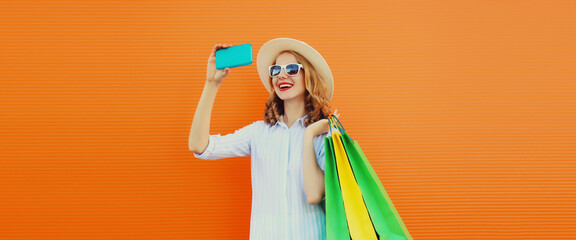 Wall Mural - beautiful happy young woman taking selfie with smartphone holding shopping bags on orange background