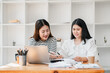 © Satori Studio - Two women in a contemporary office setting engaged in discussion over work documents with a laptop and stationery on the table.