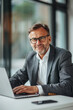 © Duka Mer - A happy businessman in a suit using a laptop at the office, sitting at a white table with glass walls in the background, wearing glasses, and looking into the camera.