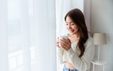 Fototapeta  - Young asian woman standing beside window and holding mug in bedroom at home, She drinking milk after wake up in the morning