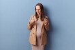 Displeased confused young adult pregnant woman wearing dress and jacket posing isolated over blue background drinking takeaway coffee and using mobile phone checking email