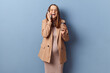 Cheerful laughing young adult pregnant woman wearing dress and jacket posing isolated over blue background talking on mobile phone expressing happiness drinking coffee